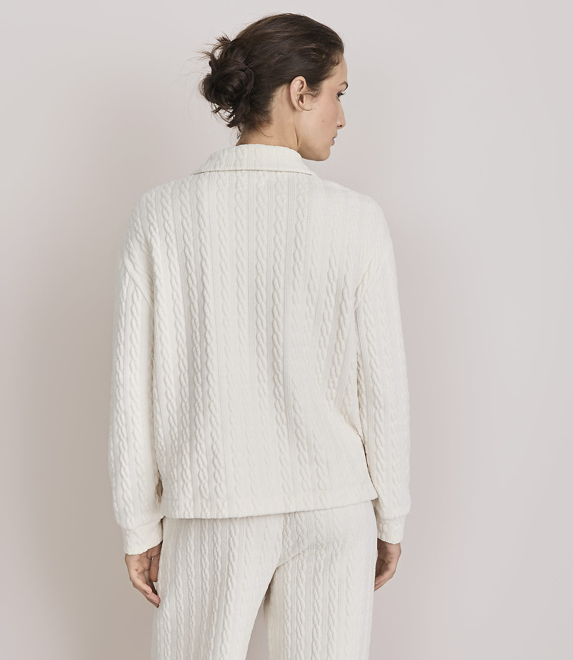 A woman in a cream cable-knit sweater and pants stands against a plain, light-colored background.