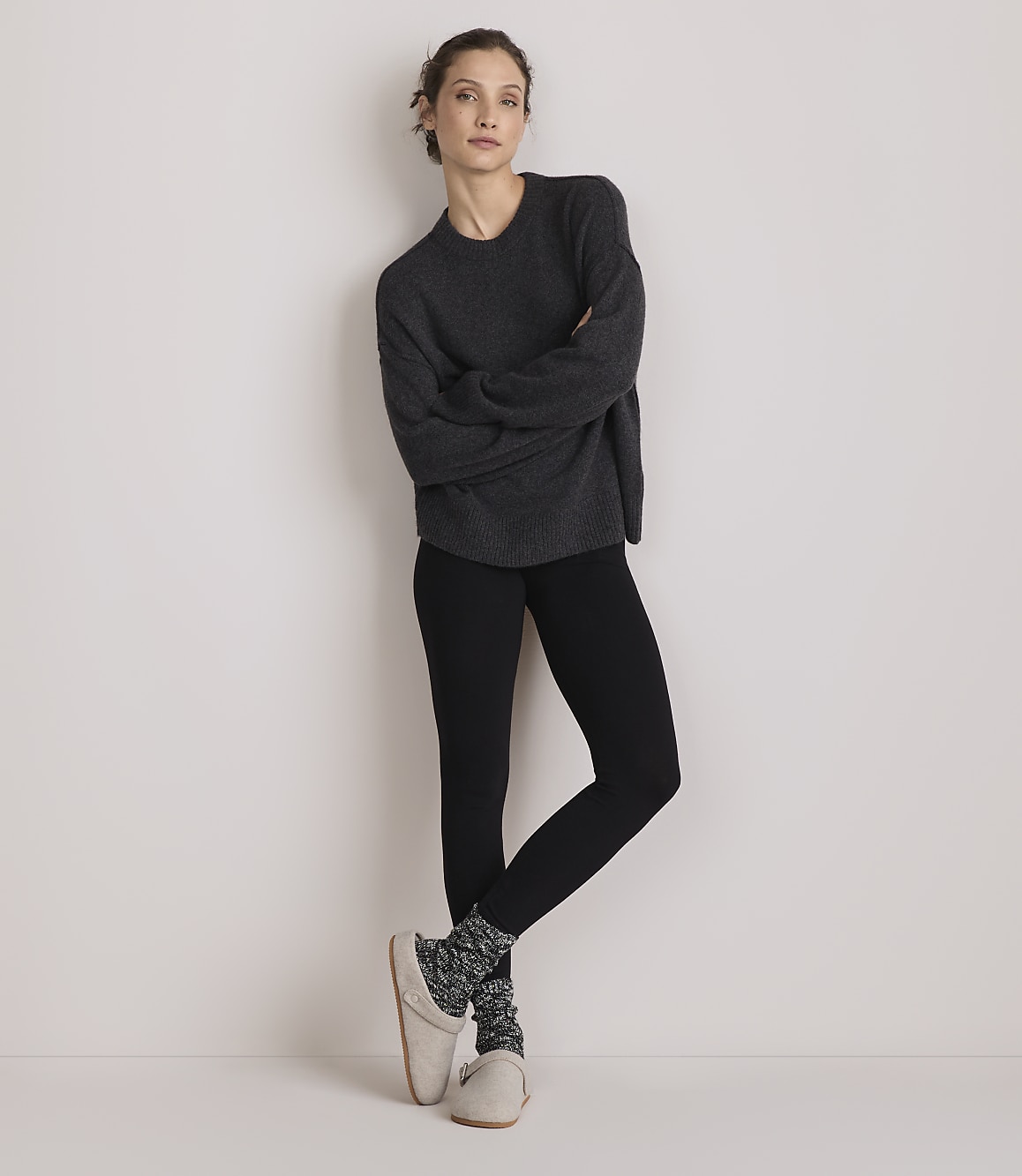 A woman in a dark gray sweater and black leggings stands with her arms crossed against a plain white background.