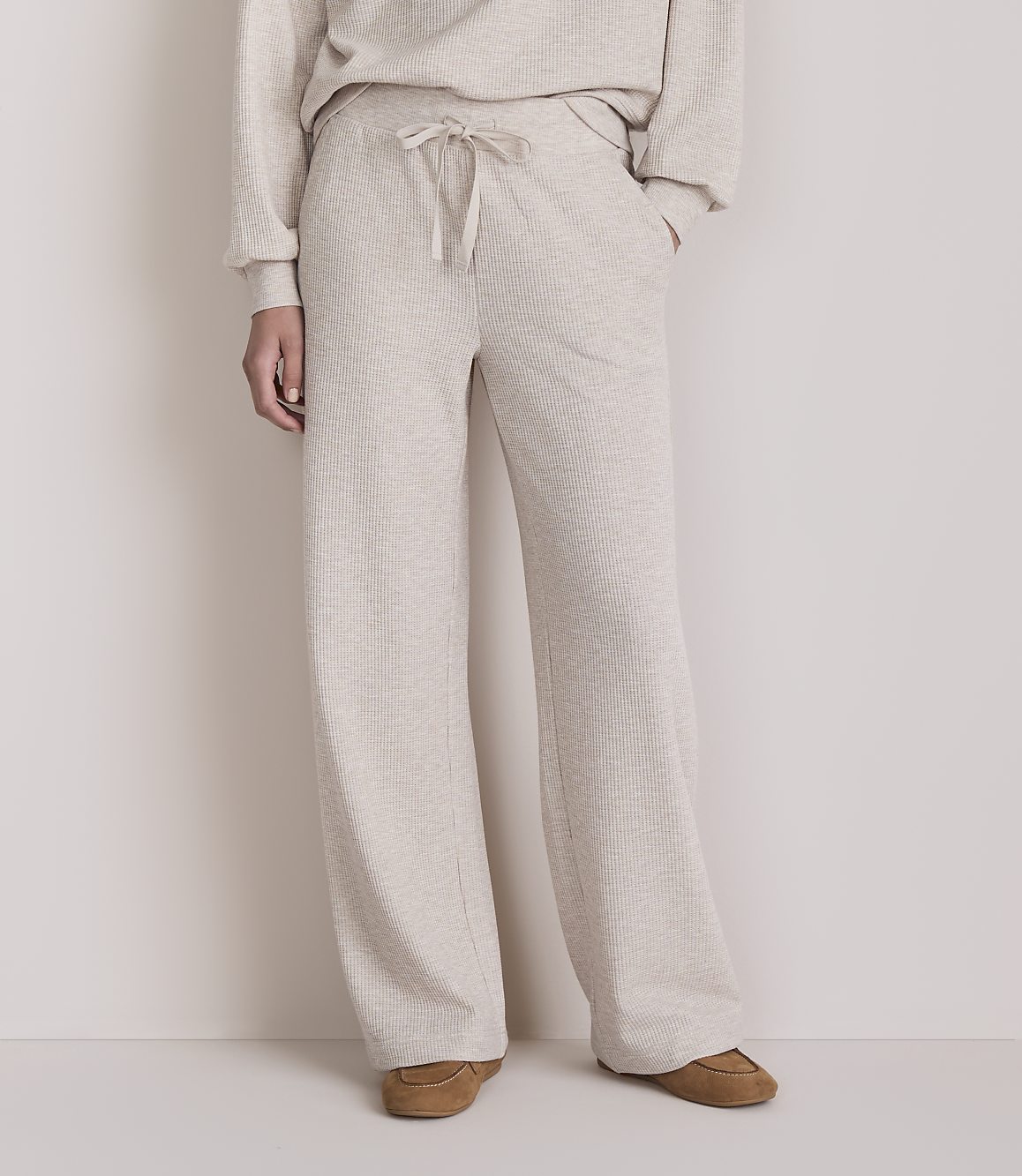 A person stands in light-colored lounge pants and slippers against a plain, light-colored background.