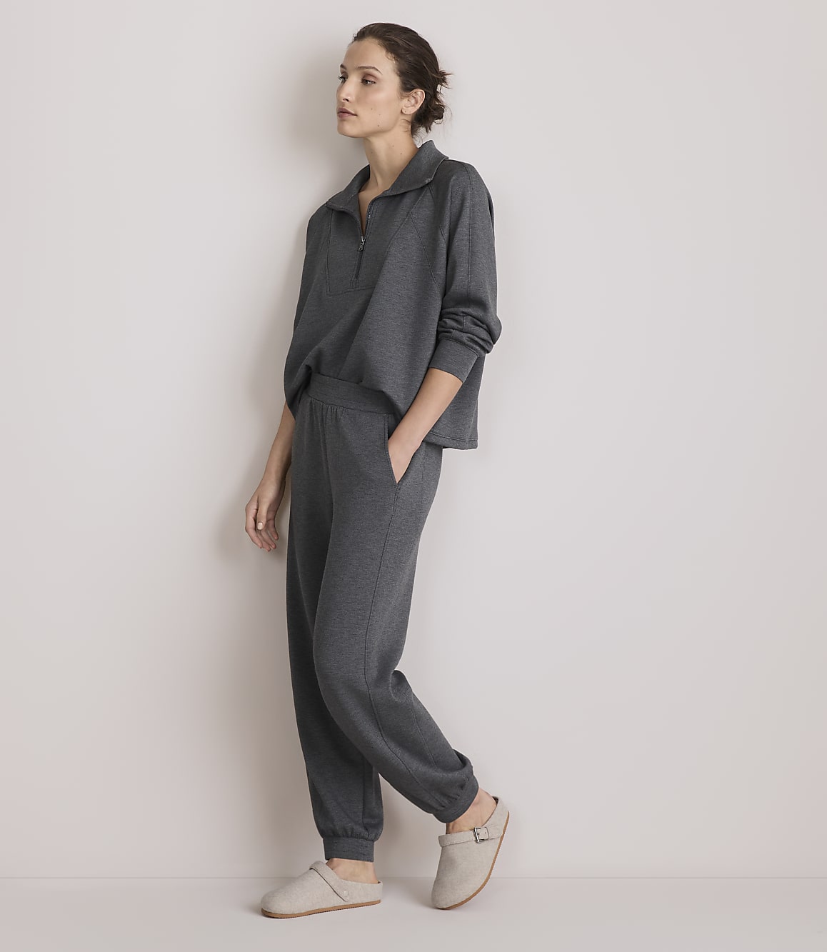 A woman in a grey loungewear set stands against a plain, light-colored wall.