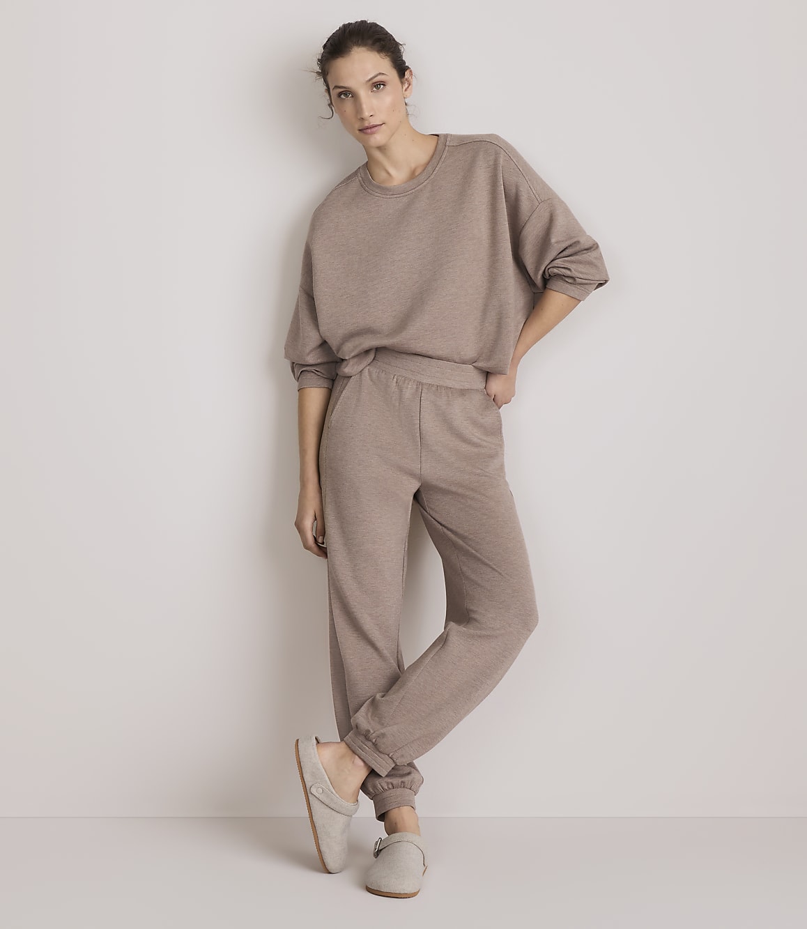 A woman in a beige loungewear set and slippers stands against a plain, light-colored wall.