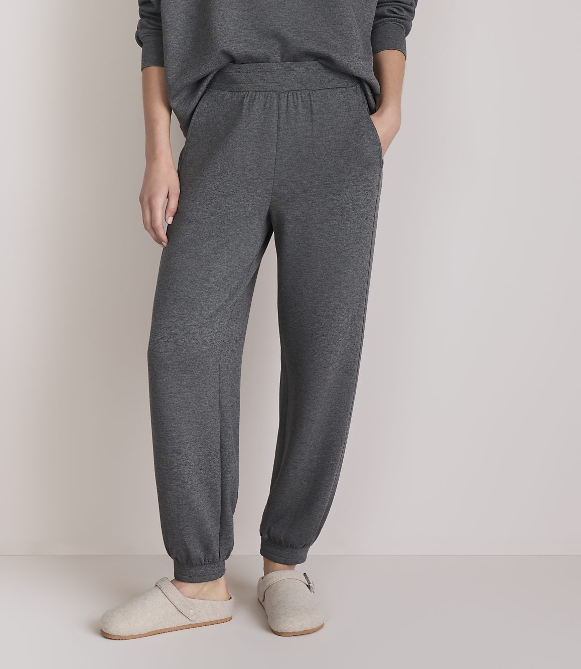 A person stands in comfortable grey loungewear and slippers against a plain, light-colored background.