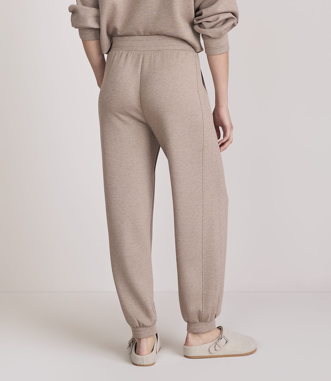 A person wearing beige sweatpants and slippers stands against a plain, light-colored background.