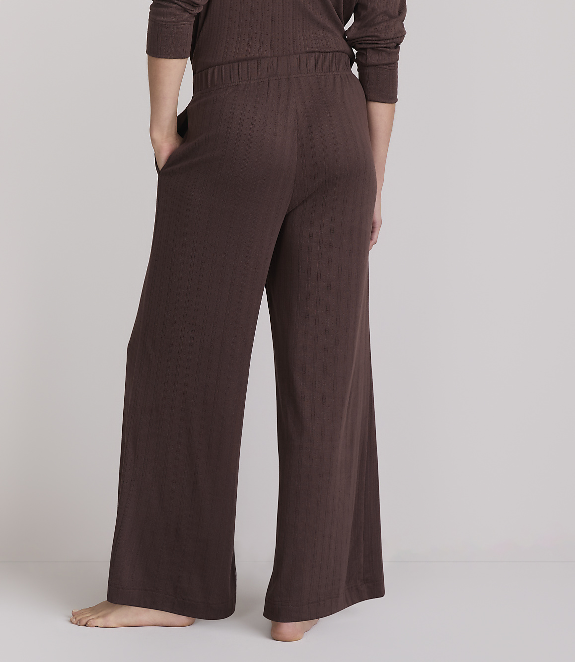 A person wearing brown ribbed pajama pants stands with their back to the camera against a plain, light gray background.