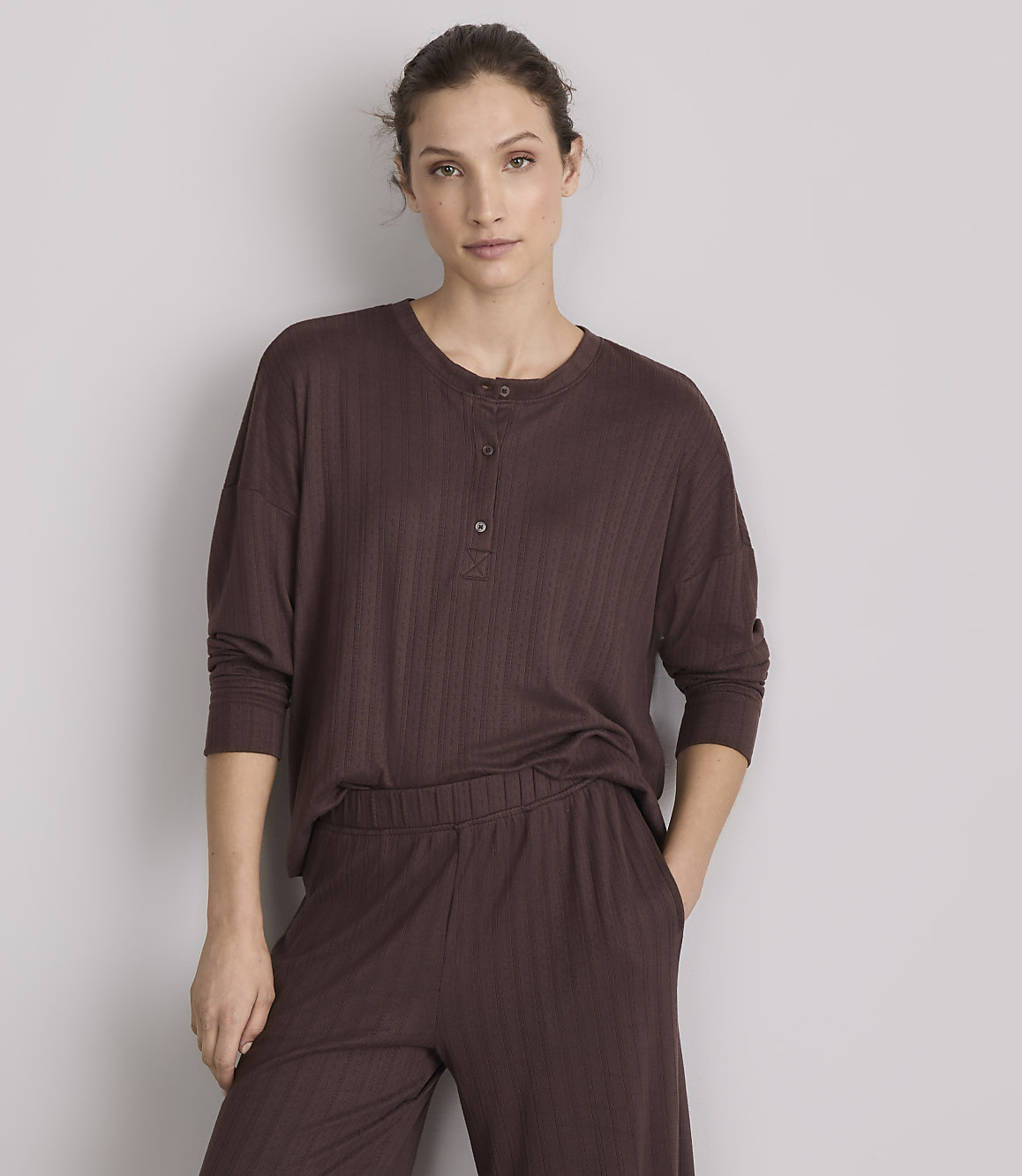 A woman in a brown ribbed henley shirt and matching pants stands against a plain gray background.