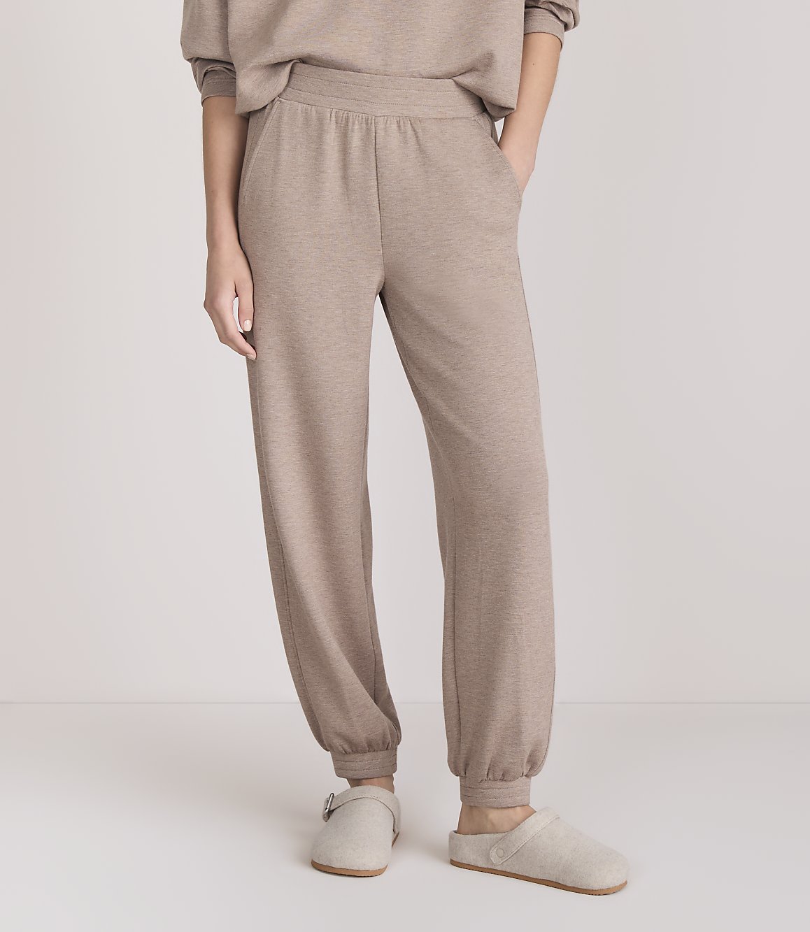 A person stands in beige loungewear and slippers against a plain white background.
