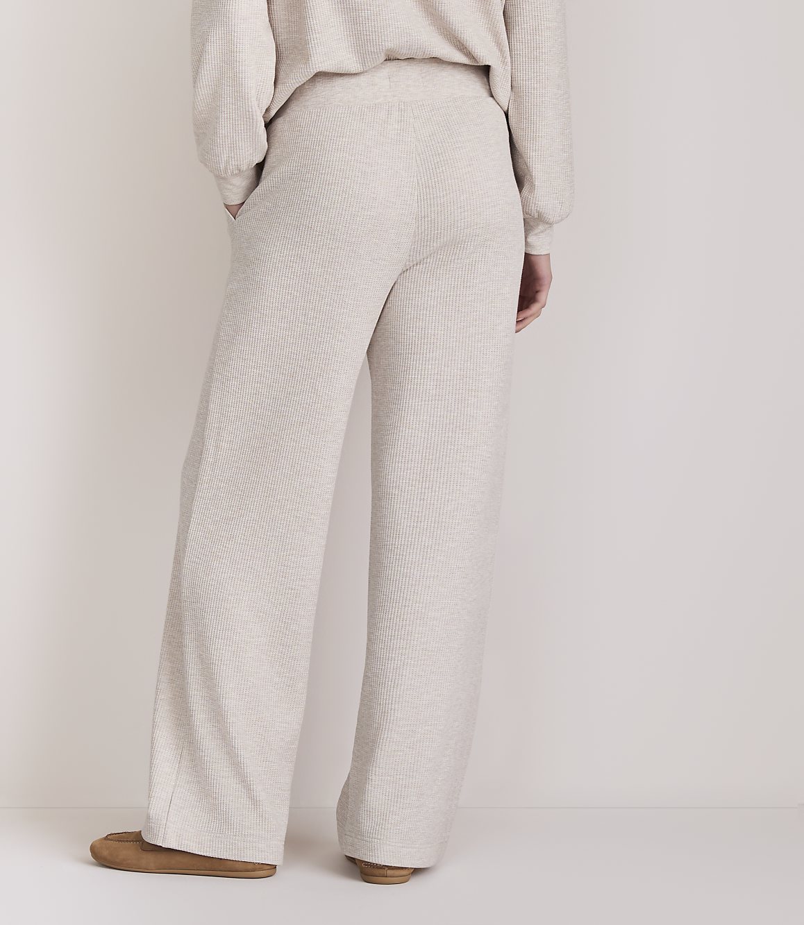 A person wearing light-colored, wide-leg lounge pants and slippers stands against a plain, light-colored background.