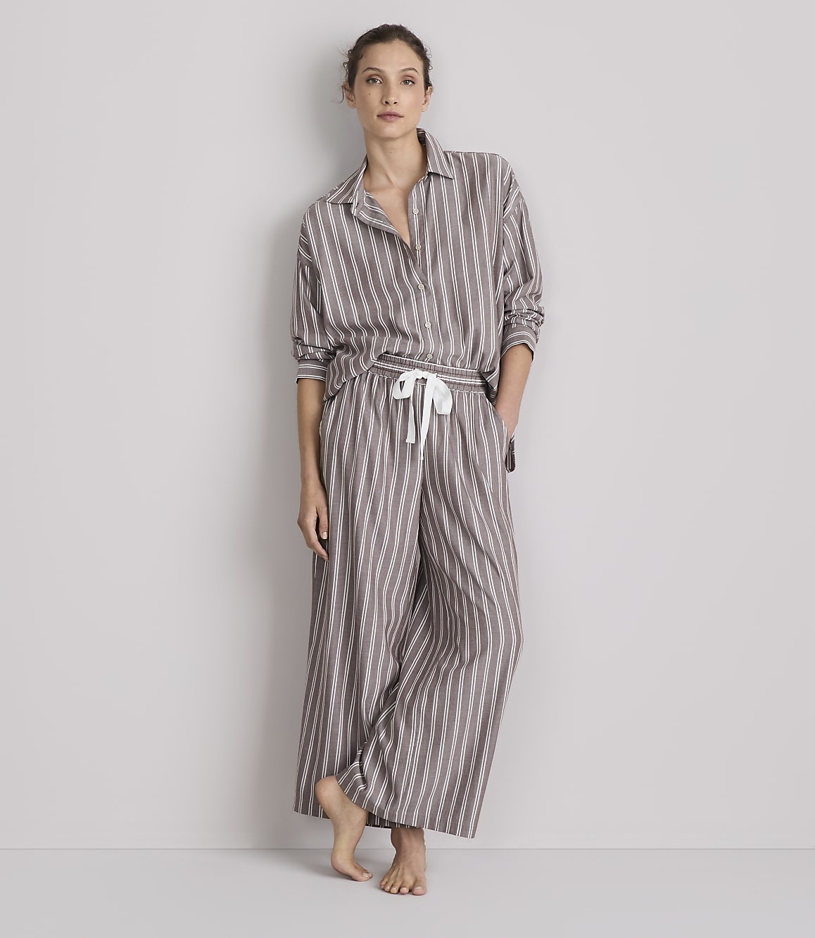 A woman stands barefoot in striped pajamas against a plain gray background.