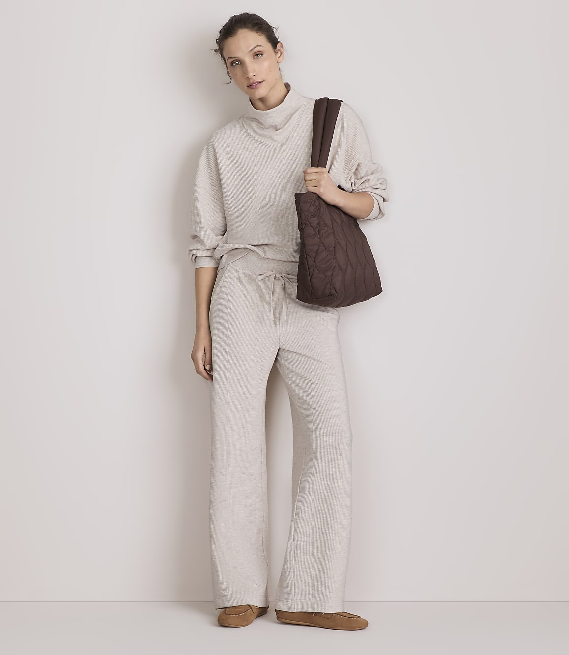 A woman in a beige loungewear set and brown tote bag stands against a plain, light-colored wall.