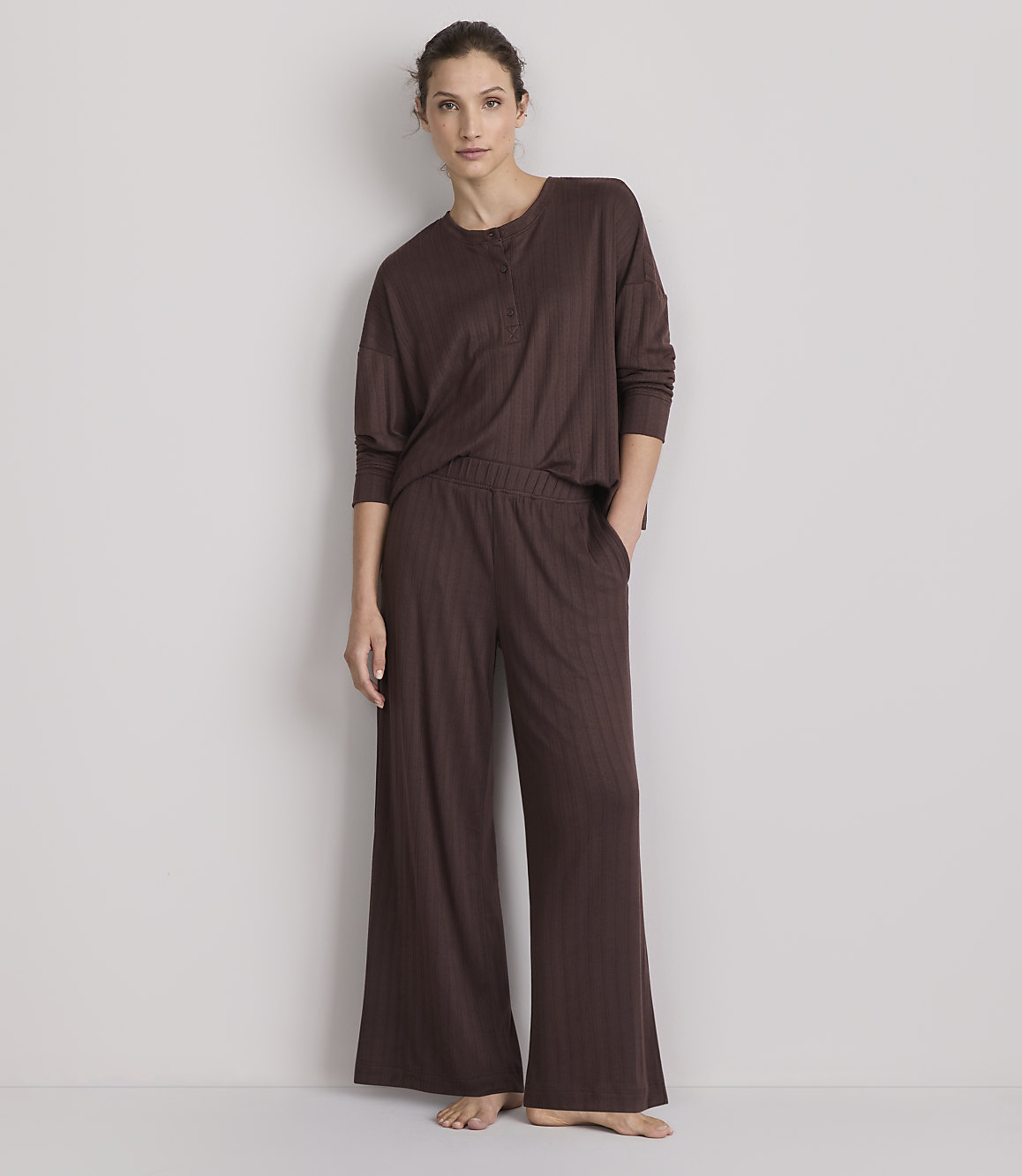 A woman in a brown ribbed pajama set stands against a plain, light-colored wall.