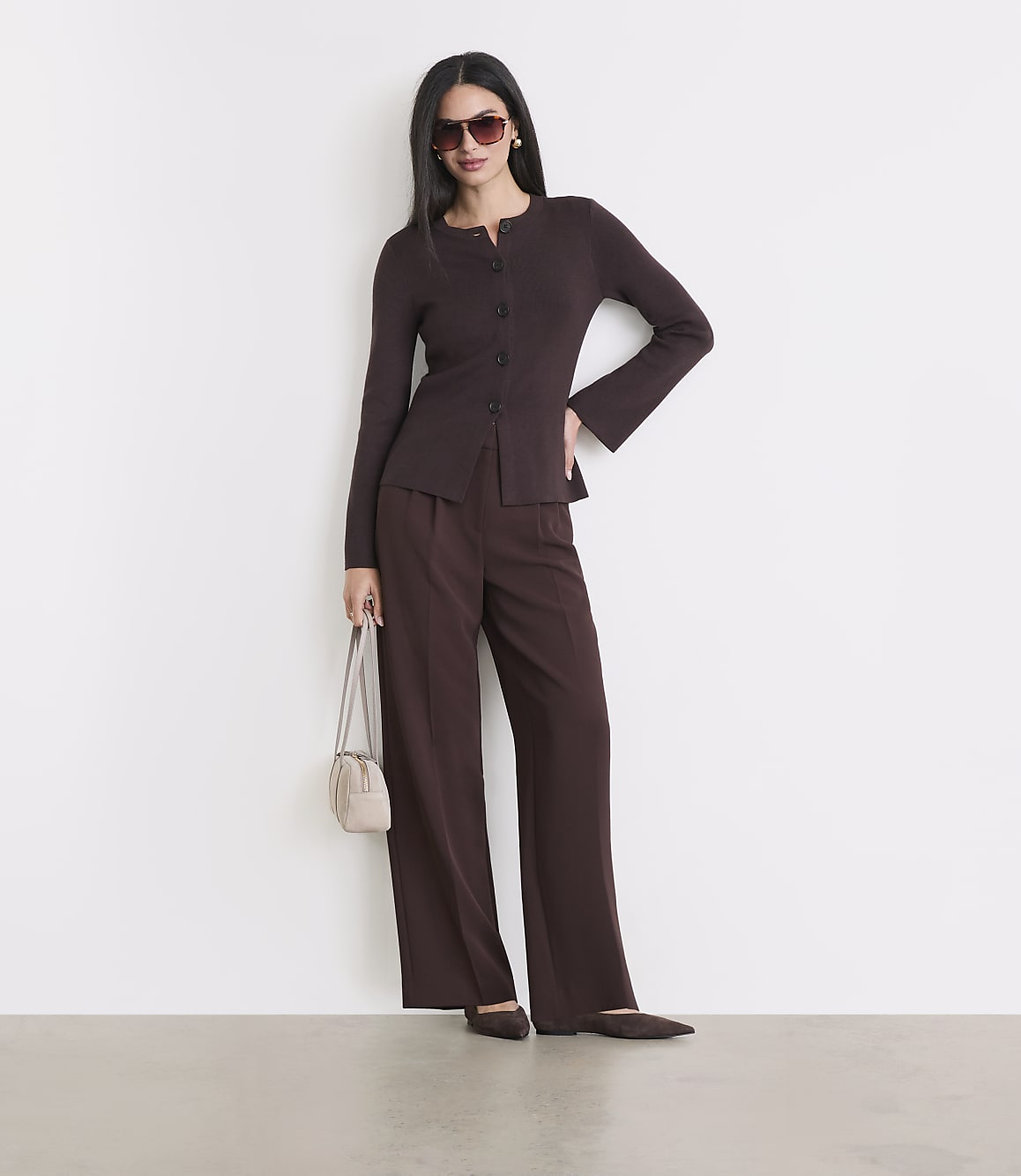 A woman in a brown cardigan and pants stands against a white wall, holding a small purse.