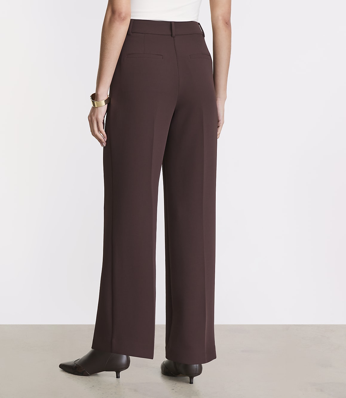 A person wearing brown wide-leg trousers and pointed-toe heels stands against a plain white background.