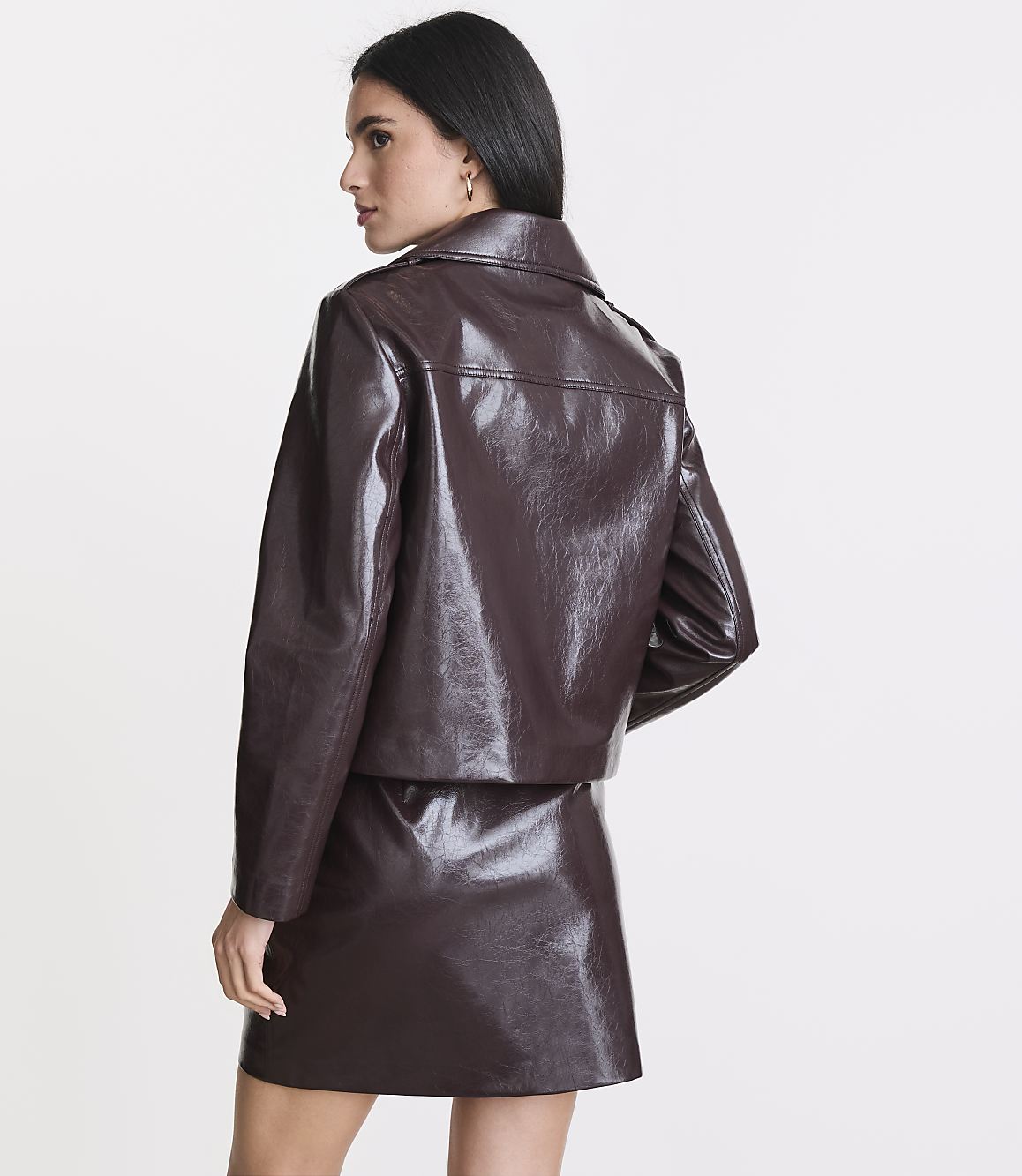 A woman in a glossy brown jacket and skirt stands against a plain white background.
