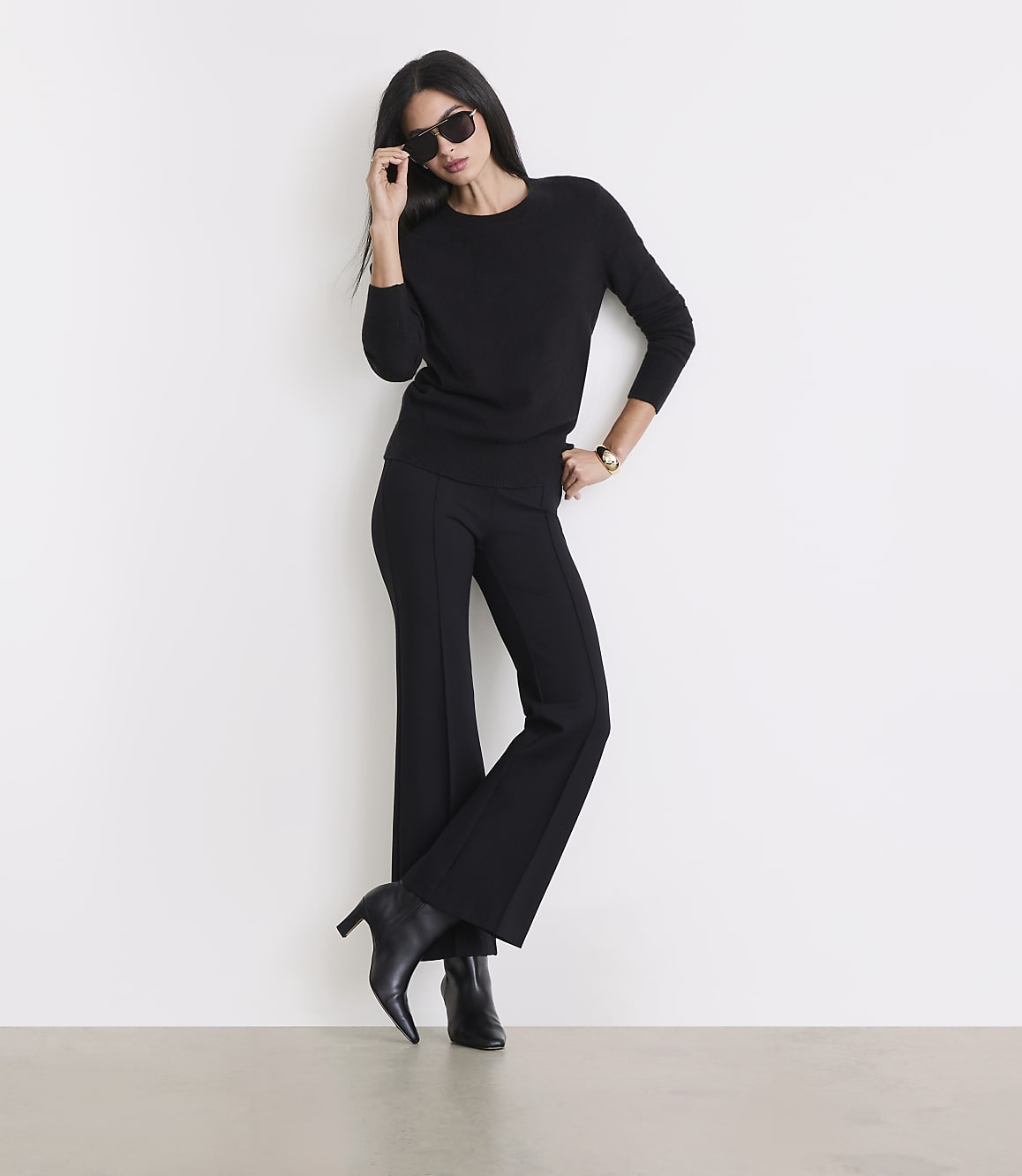 A woman in a black sweater and flared pants poses against a white wall, with a light-colored floor in the foreground.