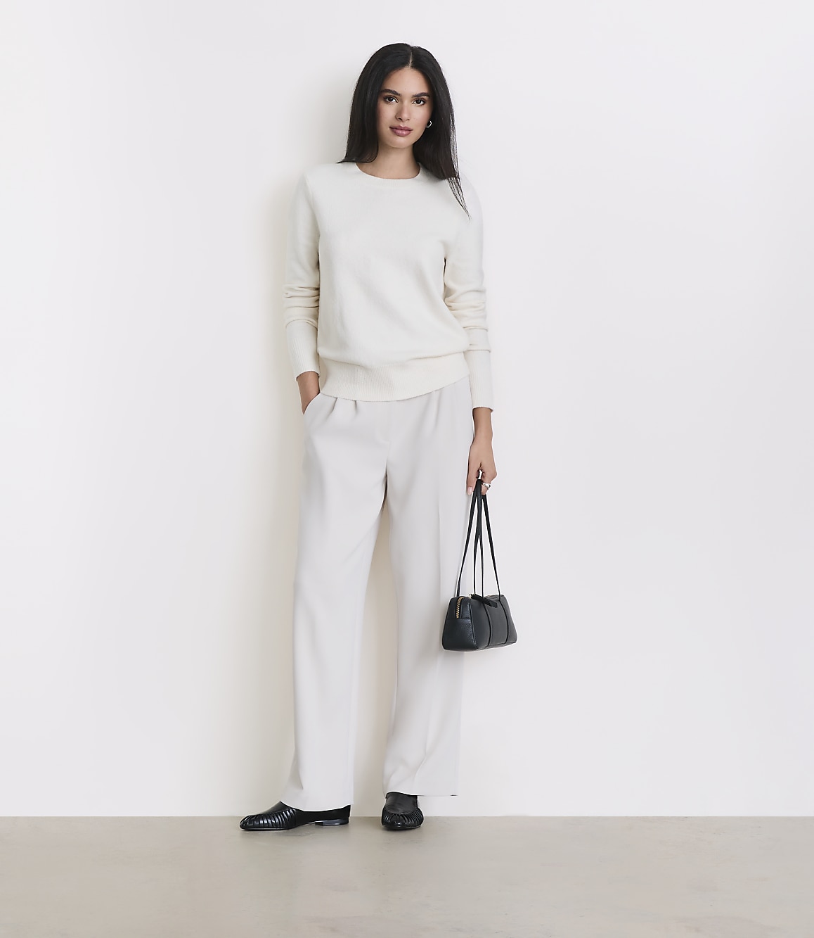 A woman in a cream sweater and matching pants stands against a plain white background, holding a small black purse.