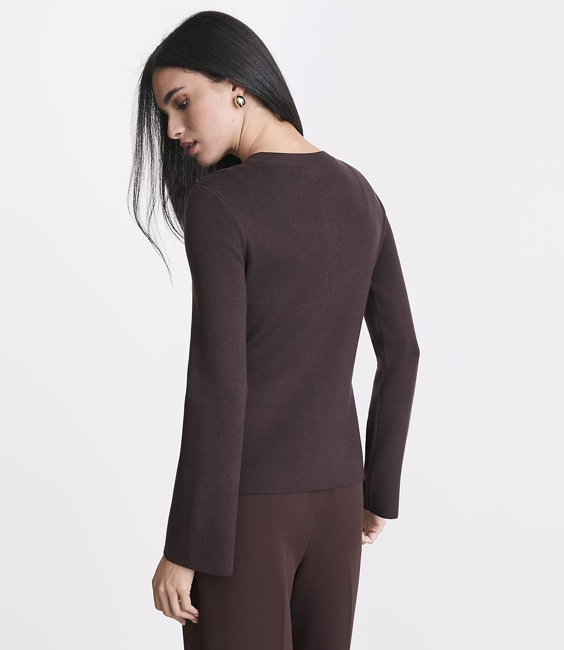A woman in a brown sweater and pants stands against a plain background.
