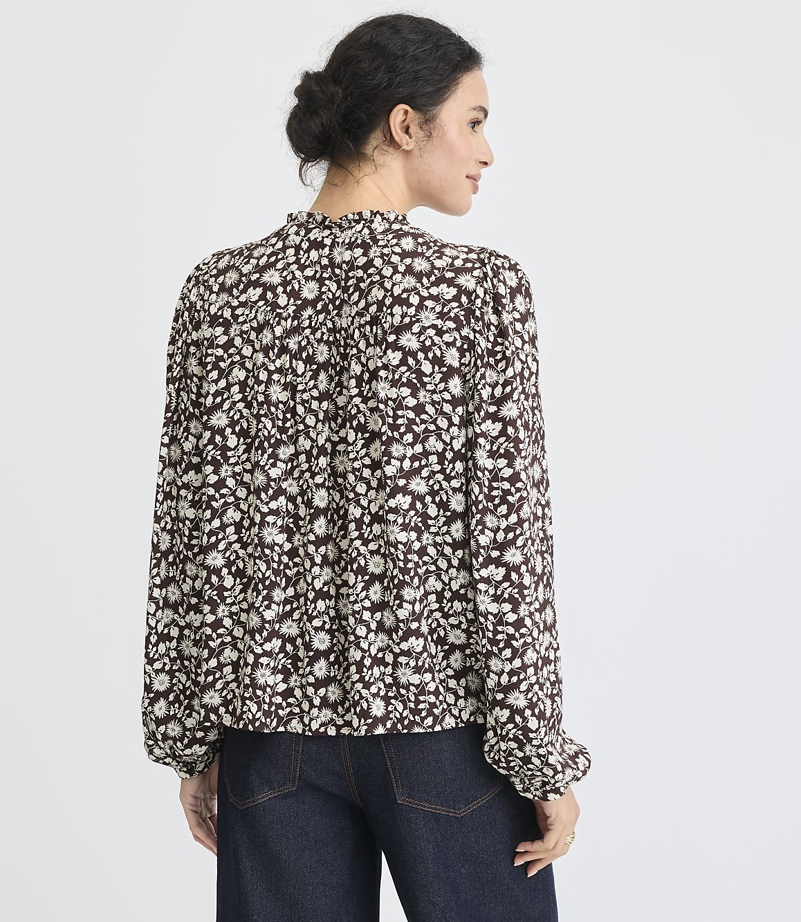 A woman in a floral blouse and dark jeans stands against a plain white background.