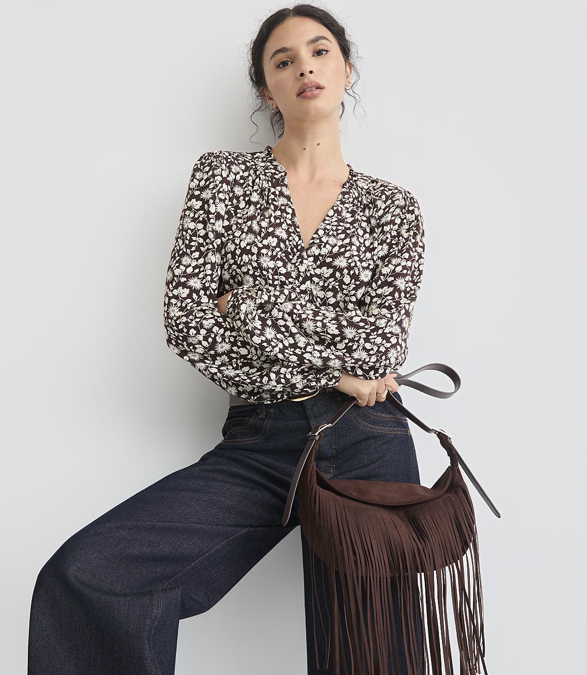 A woman in a floral blouse and dark jeans poses with a fringed suede bag against a plain white background.
