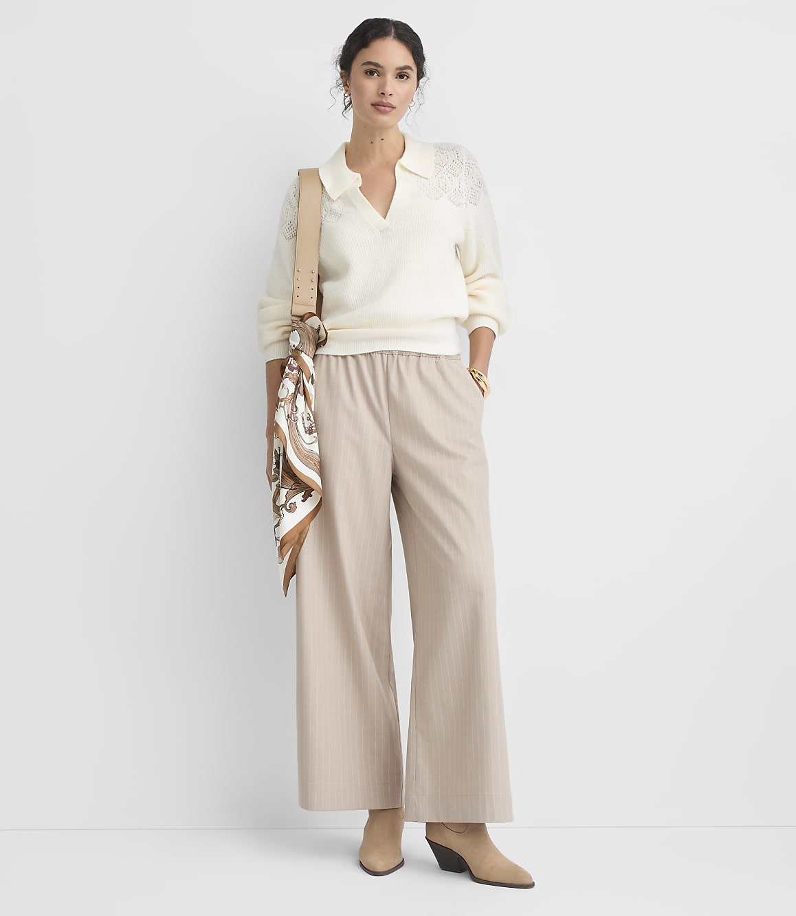 A woman in a cream collared sweater and beige pinstripe wide-leg pants stands against a plain white background, accessorized with a patterned scarf and a crossbody bag.