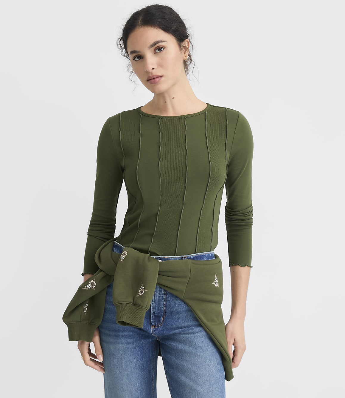 A young woman in a green ribbed long-sleeved shirt and jeans stands against a plain white background, with a green sweatshirt tied around her waist.