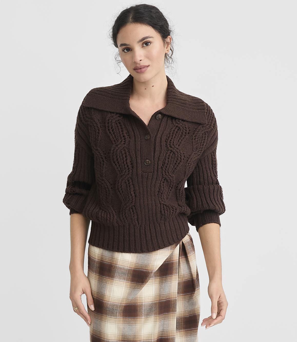 A woman in a brown cable-knit sweater and plaid skirt stands against a plain white background.