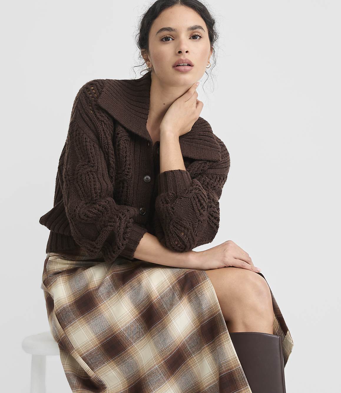 A woman in a brown cable-knit cardigan and plaid skirt sits against a plain white background.