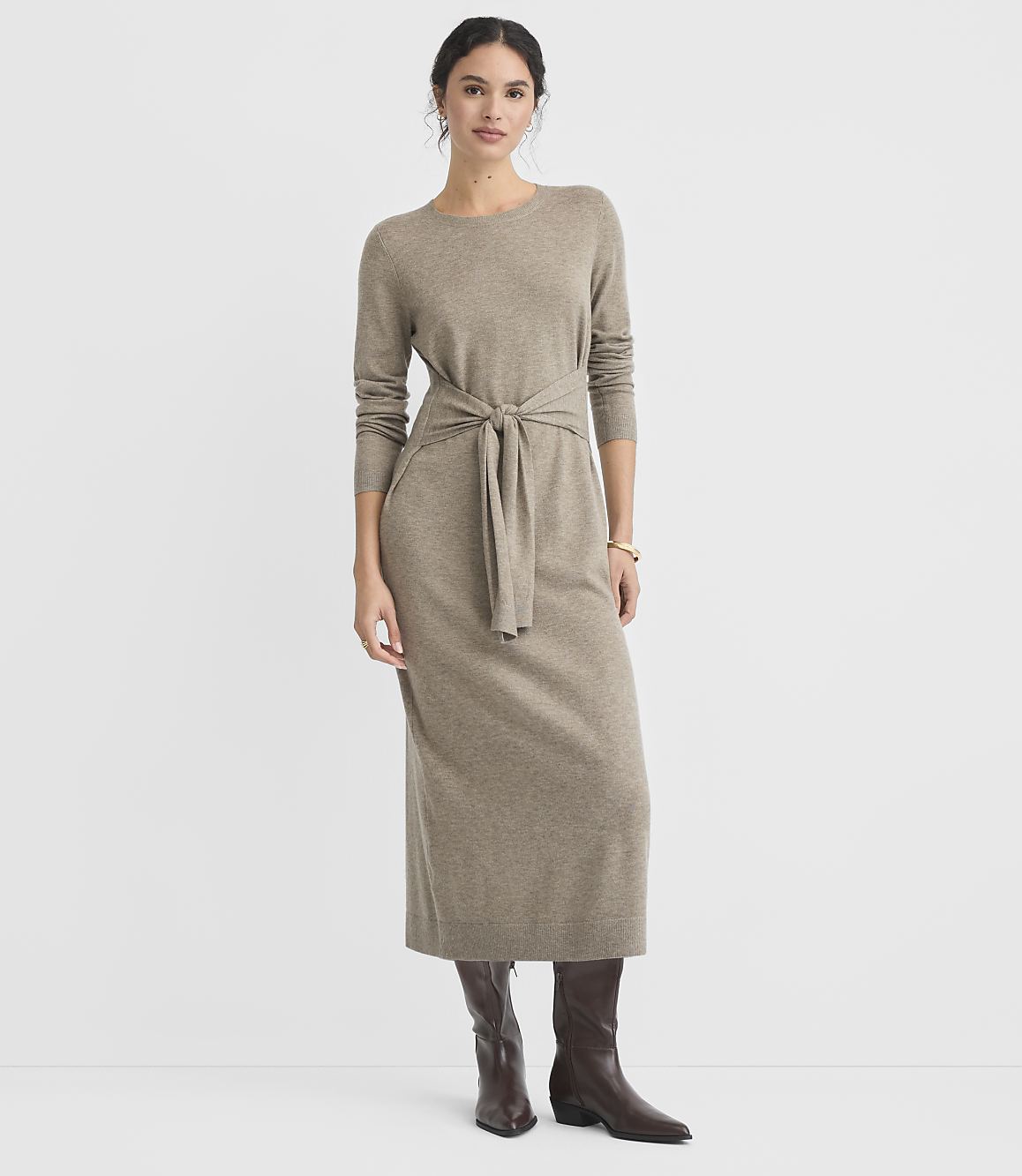 A woman in a taupe knit dress and brown boots stands against a plain white background.