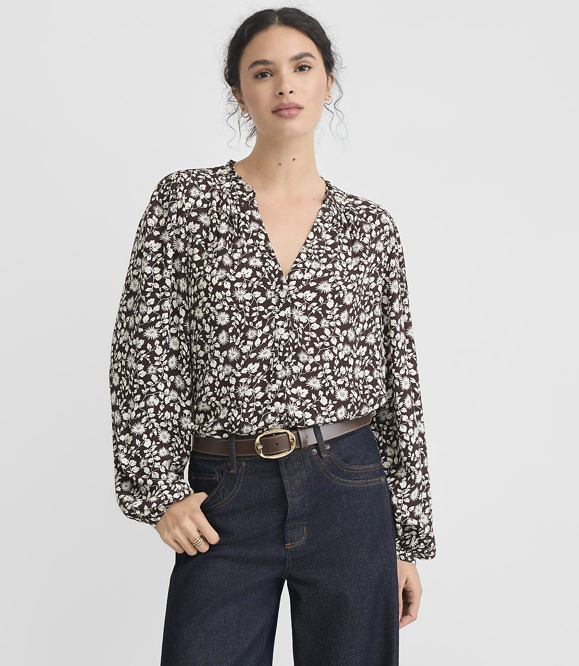 A woman in a floral blouse and jeans stands against a plain white background.