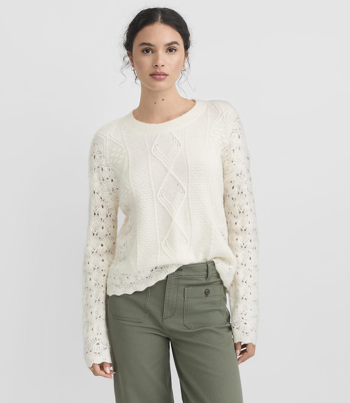 A woman in a cream cable-knit sweater and green pants stands against a plain white background.