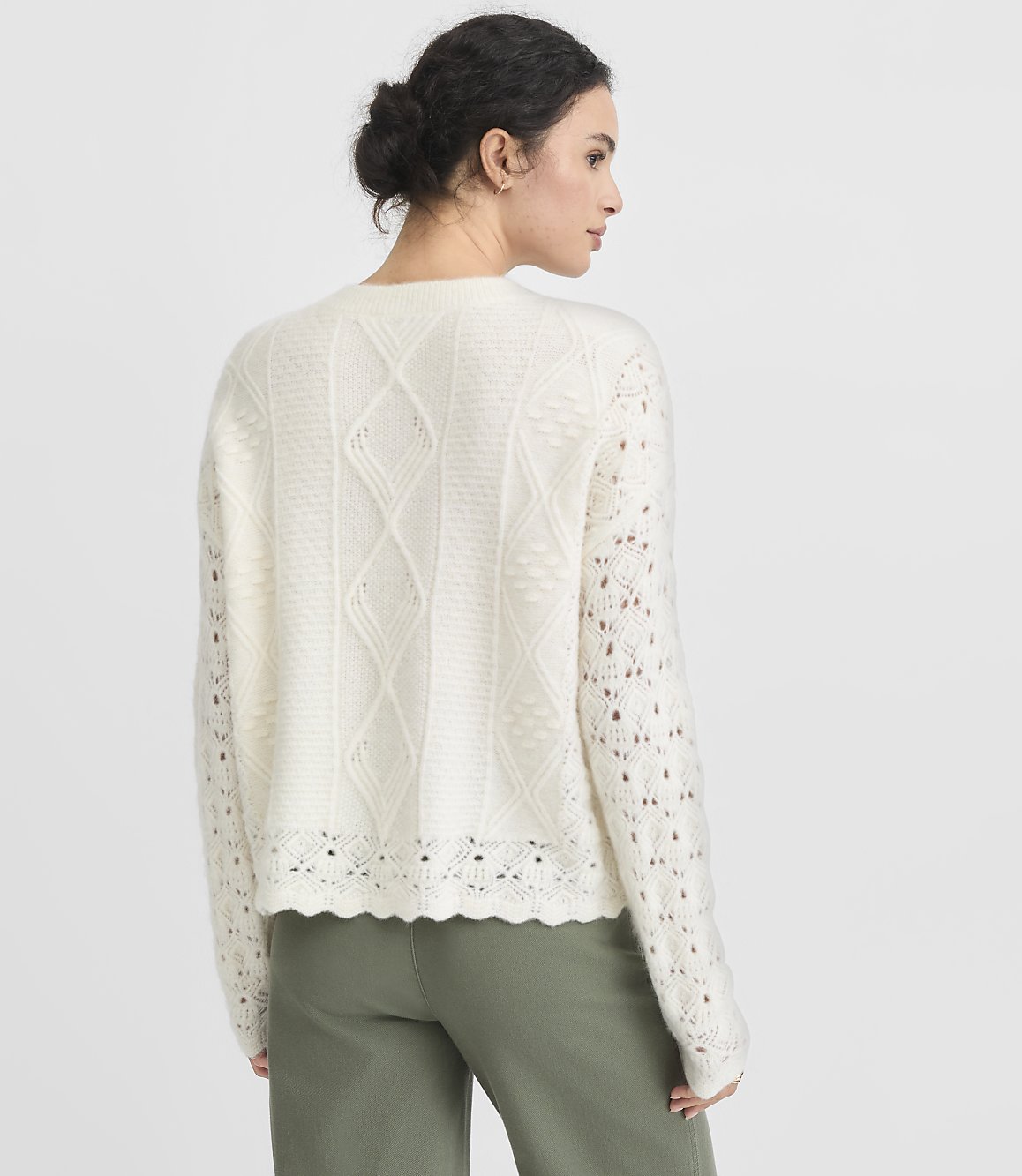 A woman in a cream-colored, cable-knit sweater and green pants stands against a plain gray background.