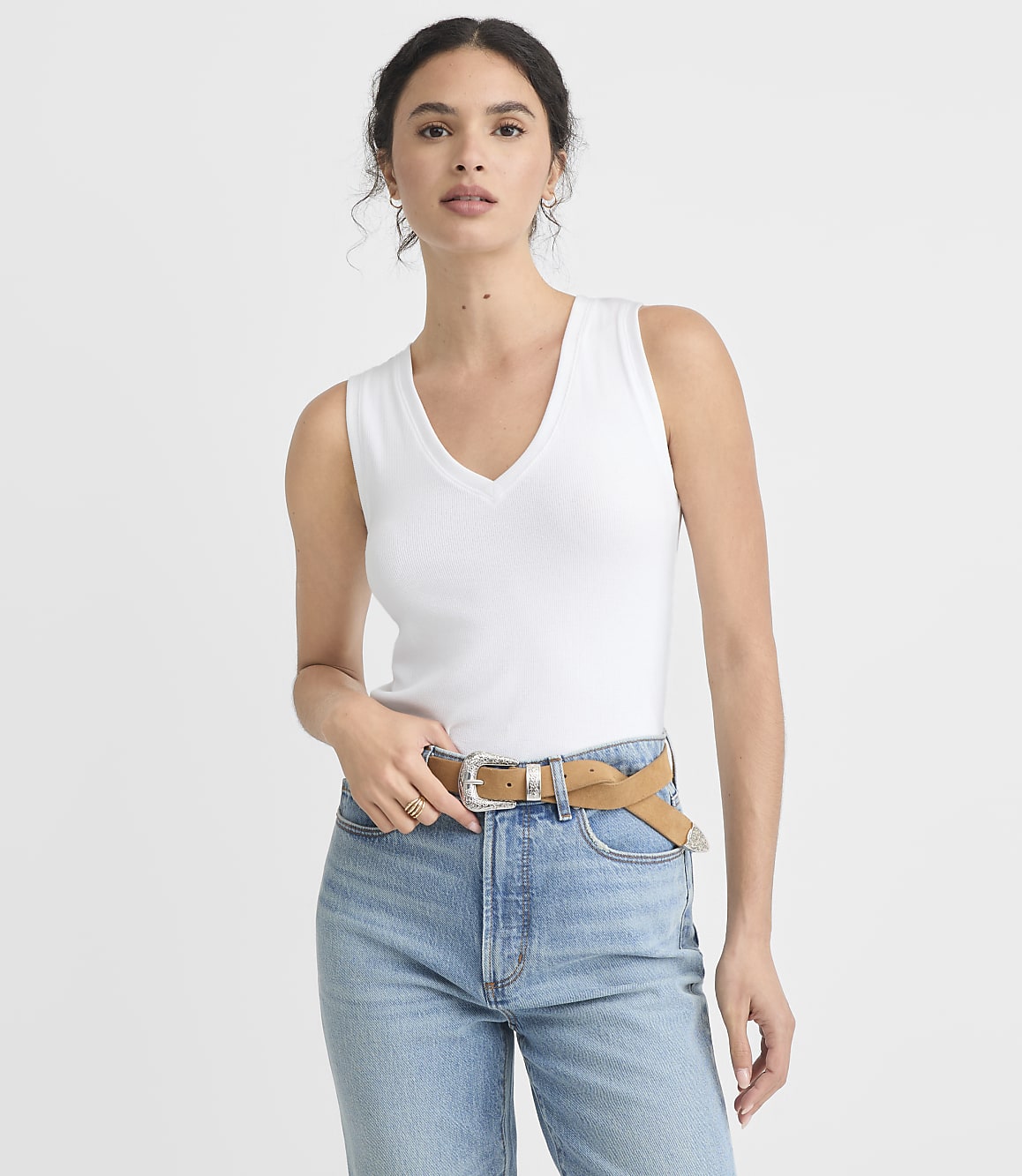 A woman in a white tank top and blue jeans with a tan belt stands against a plain white background.