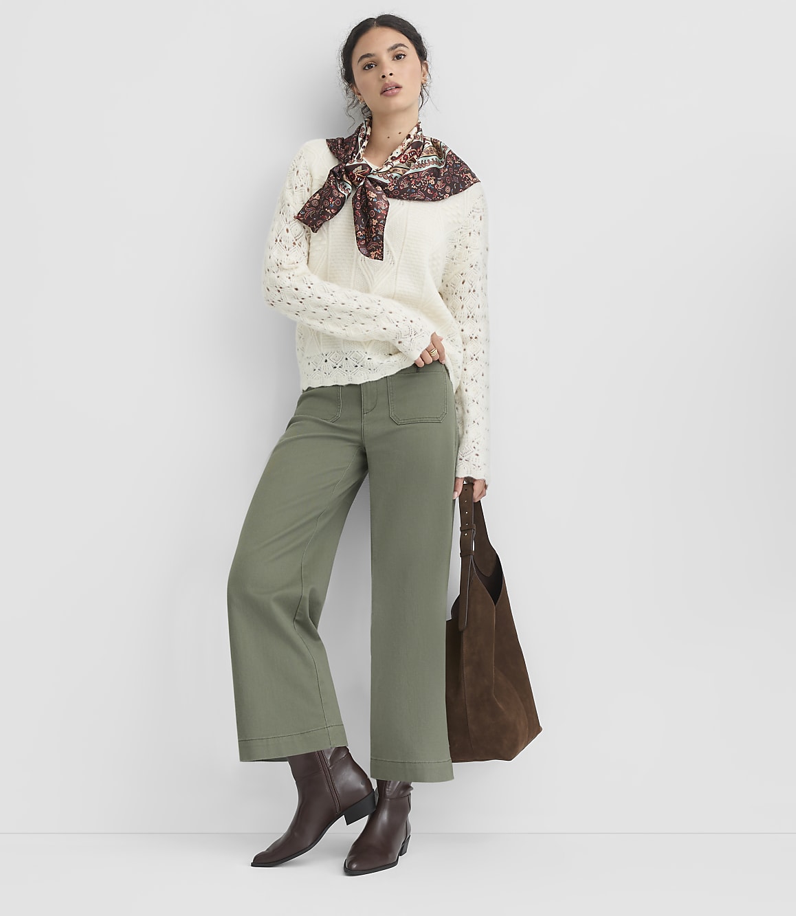 A woman in a cream sweater, olive green pants, and brown boots stands against a plain white background, holding a brown tote bag.