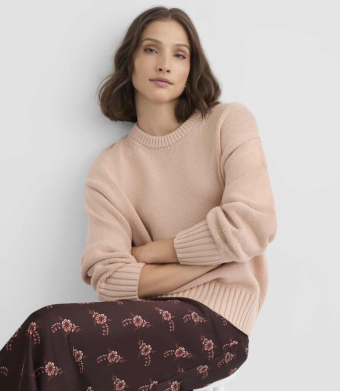 A woman in a light pink sweater and a floral skirt sits with her arms crossed against a plain white background.