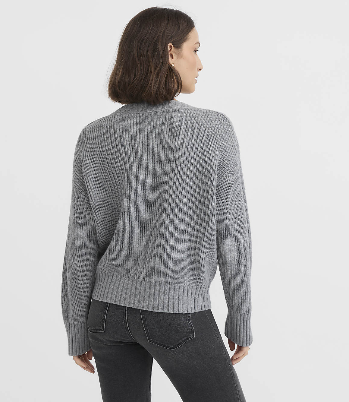 A woman in a grey sweater and jeans stands against a plain grey background.