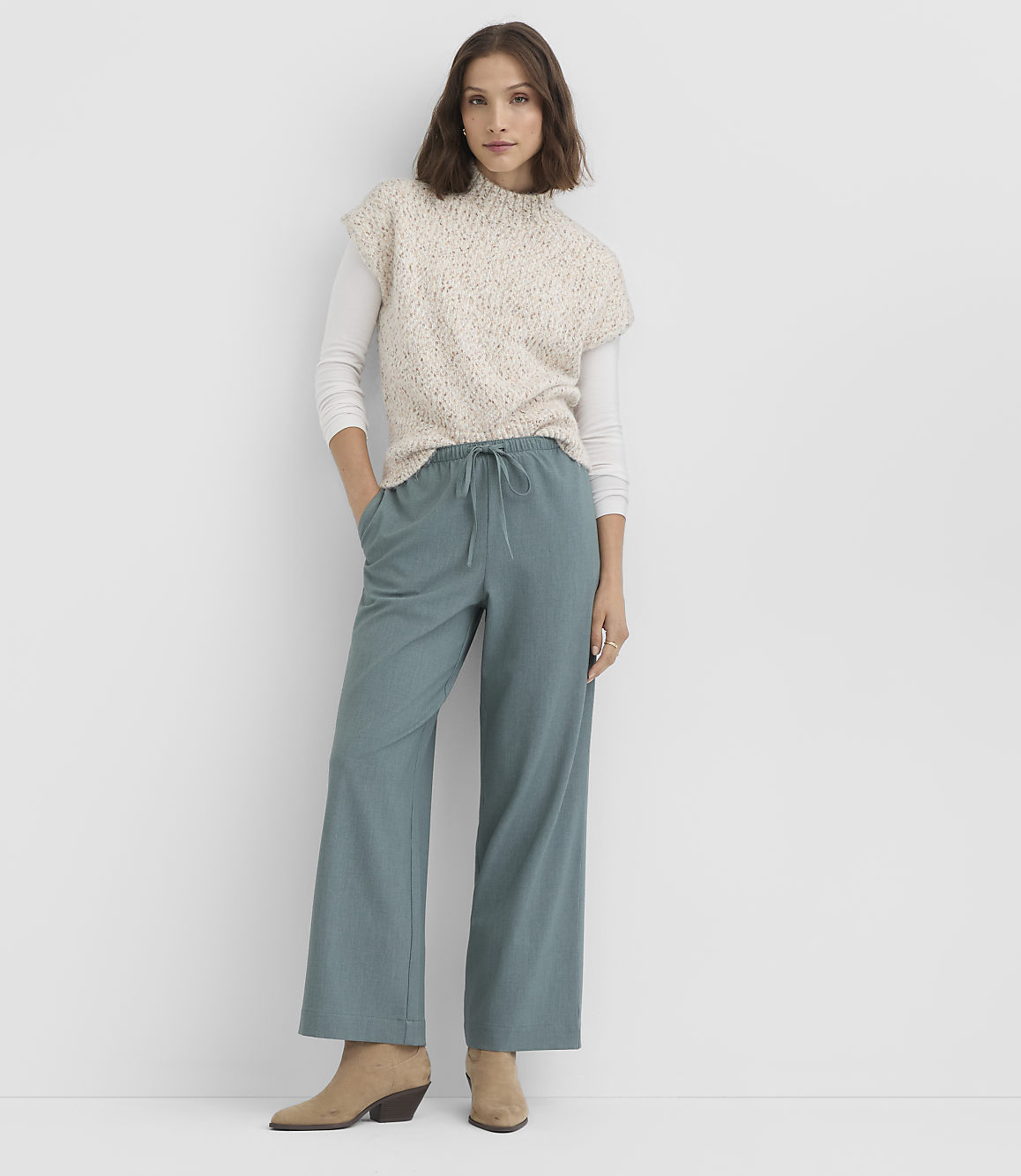 A woman in a speckled sweater vest and teal pants stands against a plain white background.