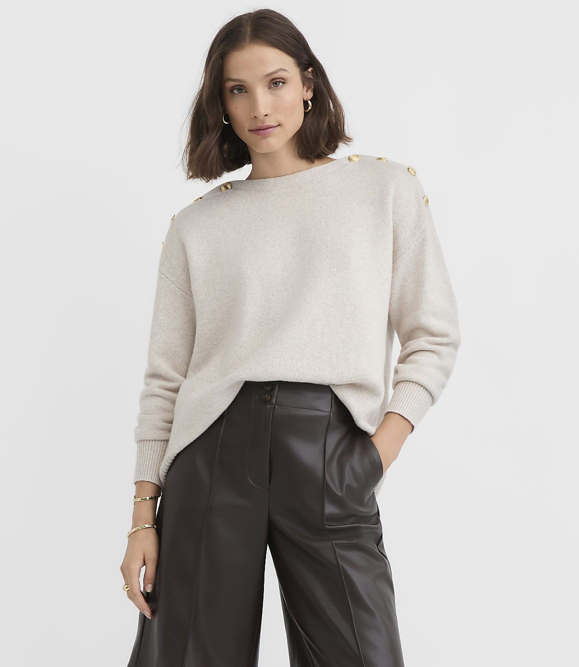 A woman in a cream sweater with gold buttons on the shoulders and dark brown leather pants stands against a plain white background.