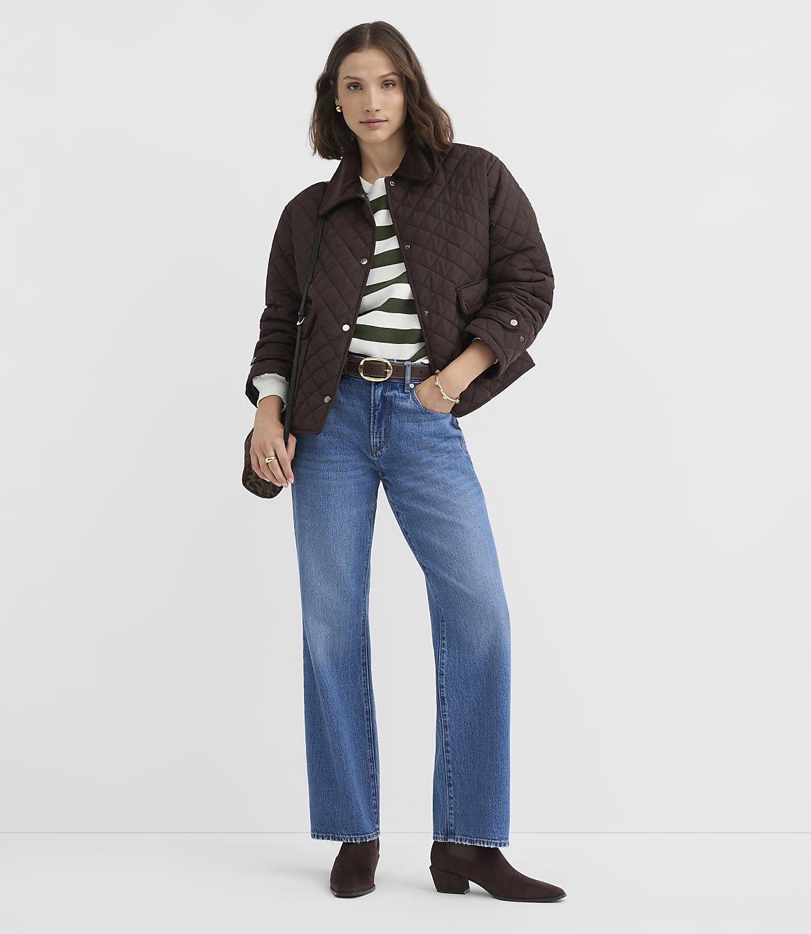 A woman in a quilted brown jacket, striped sweater, and wide-leg jeans stands against a plain gray background.