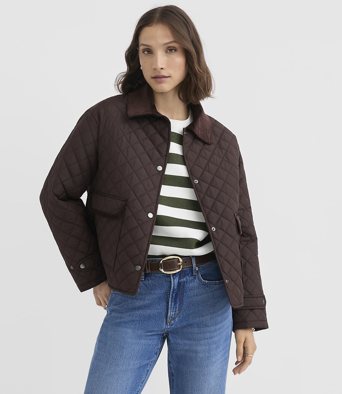 A woman in a brown quilted jacket and striped shirt stands against a plain white background.