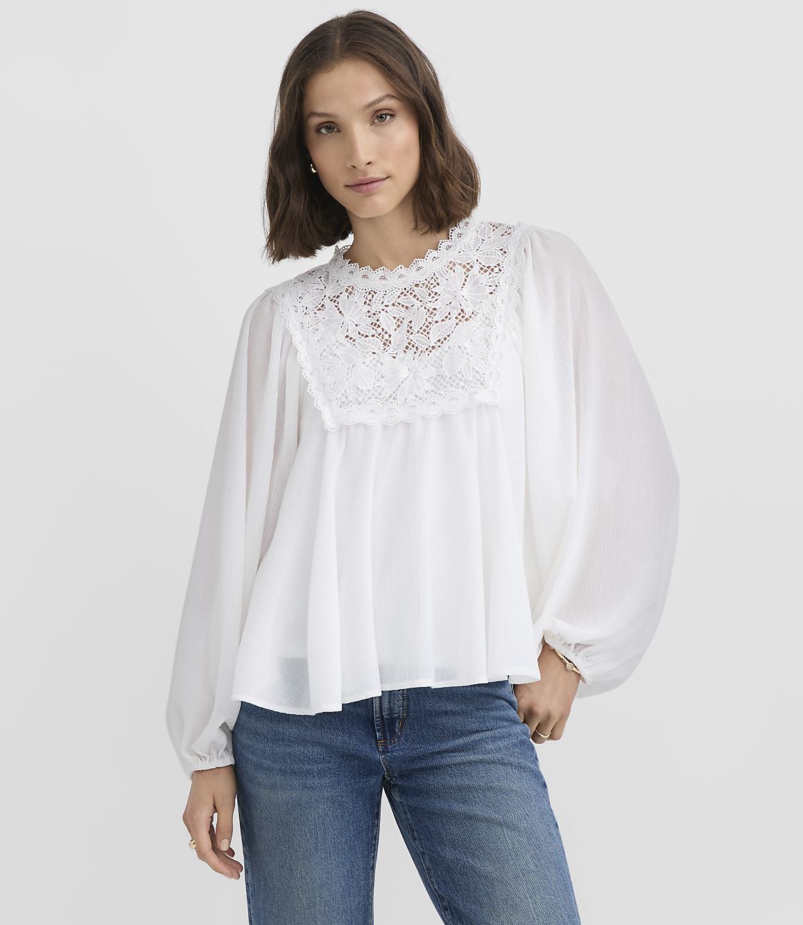 A woman in a white lace-trimmed blouse and blue jeans stands against a plain white background.