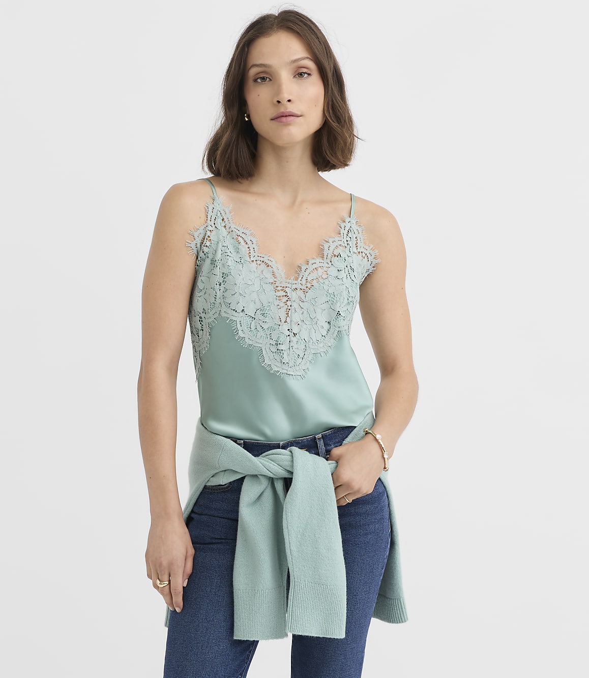 A woman in a mint green lace camisole and jeans stands against a plain white background, with a matching sweater tied around her waist.