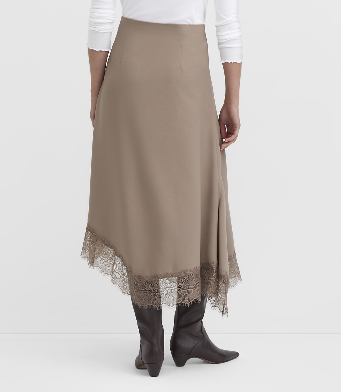 A woman in a taupe midi skirt with lace trim and brown boots stands against a plain white background.