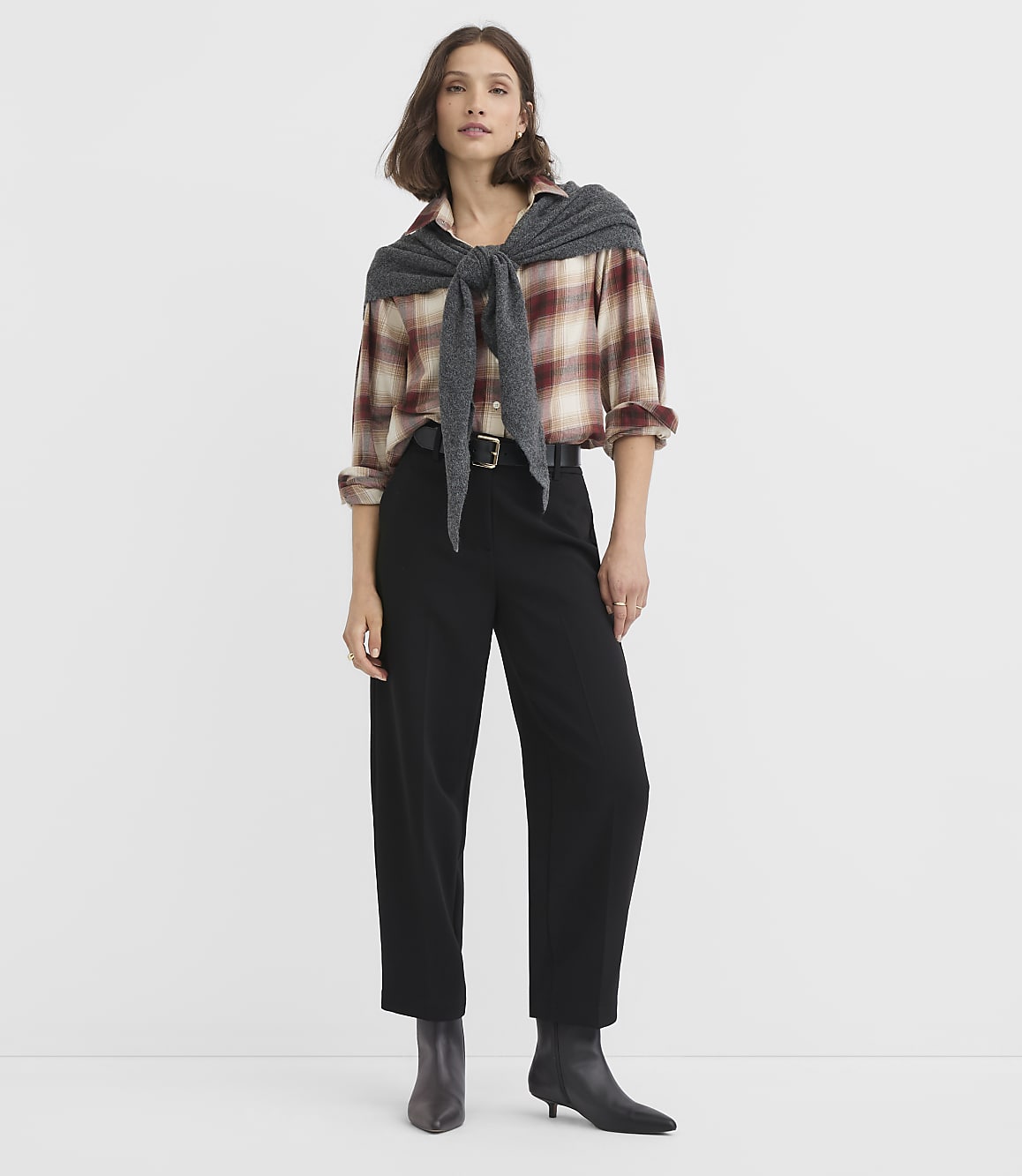 A woman in a plaid shirt and black pants stands against a plain white background.