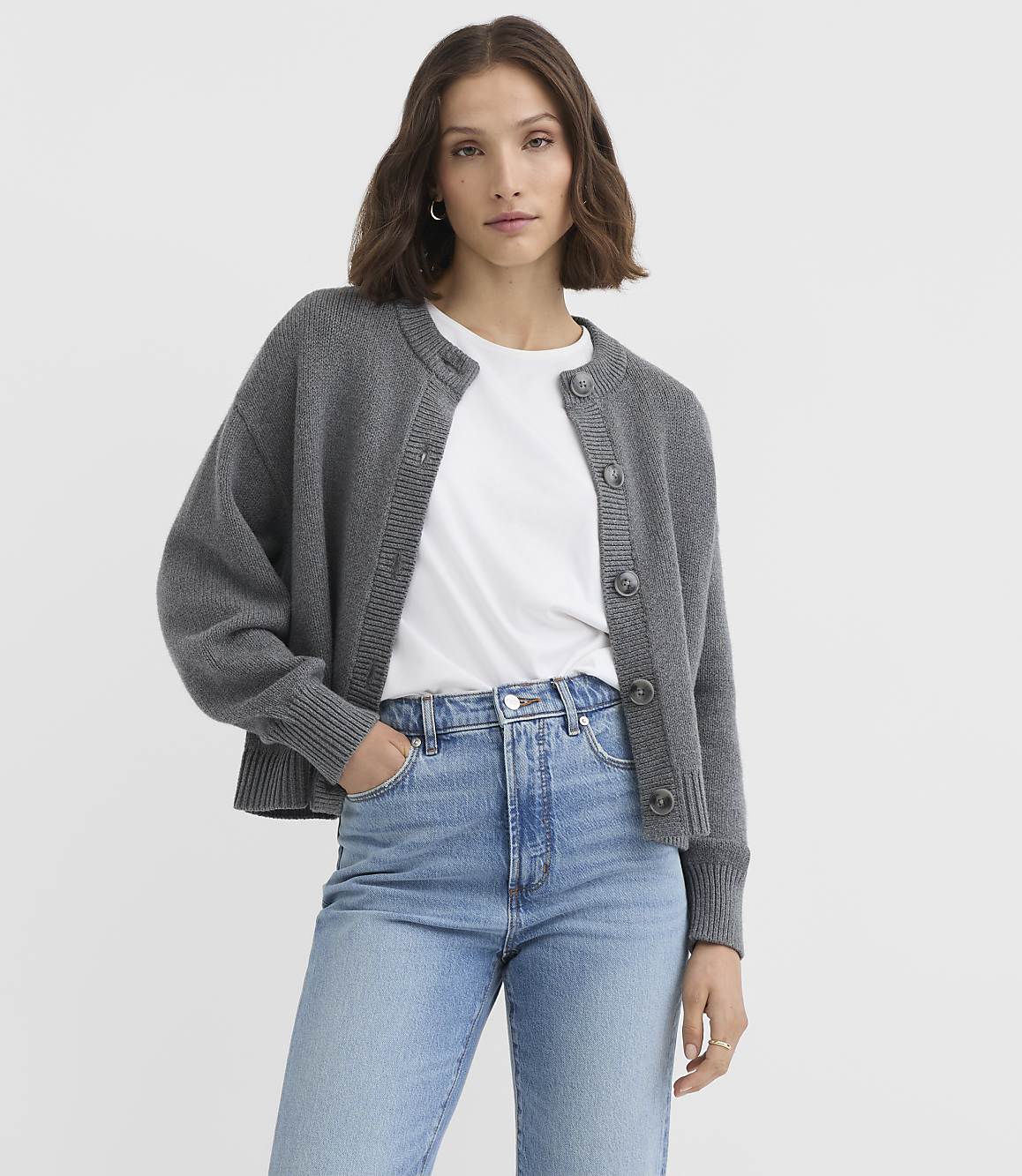 A woman in a grey cardigan and blue jeans stands against a plain grey background.