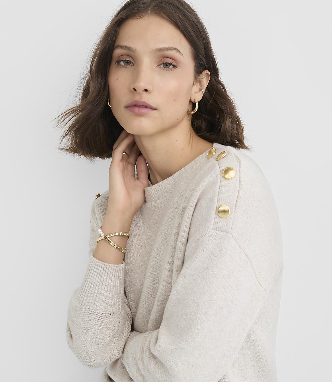 A woman in a cream-colored sweater with gold buttons on the shoulder poses against a plain gray background.