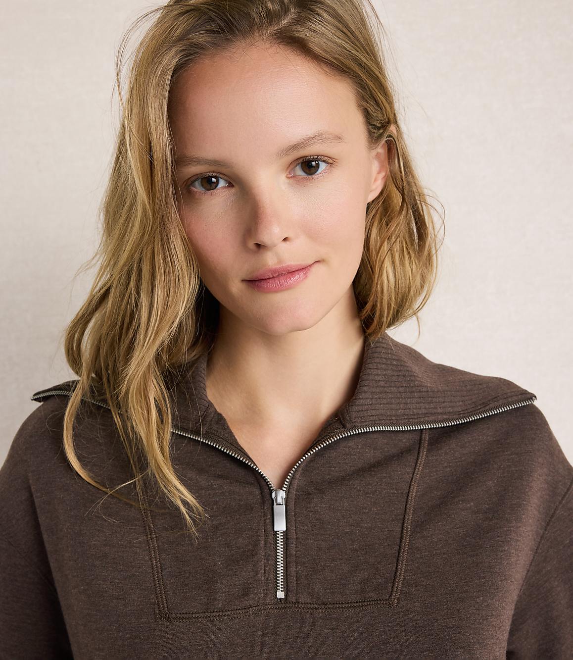 A young woman with blonde hair and a brown zippered sweatshirt is shown against a textured, light-colored background.