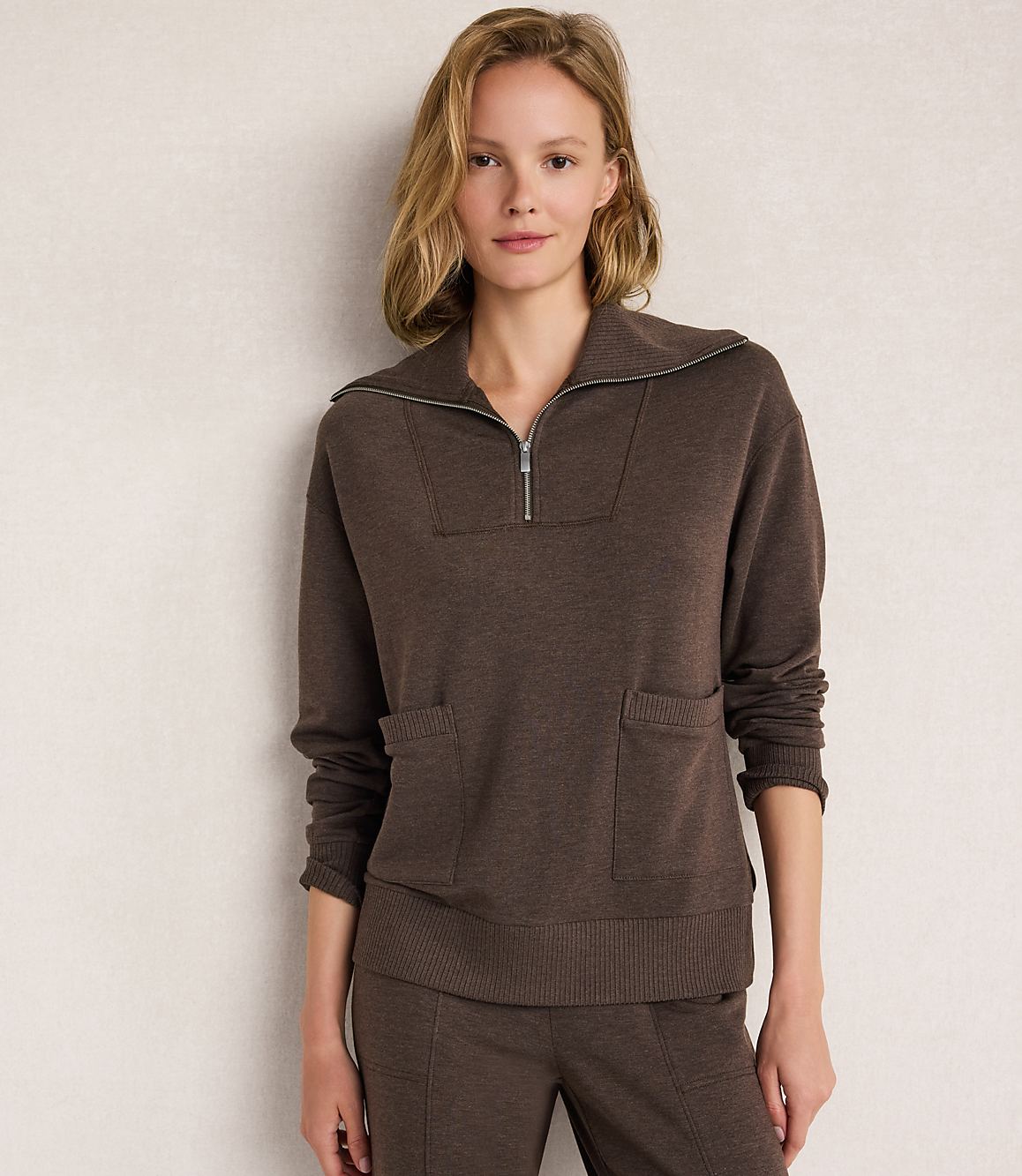 A woman in a brown quarter-zip sweater and matching pants stands against a plain, light-colored wall.