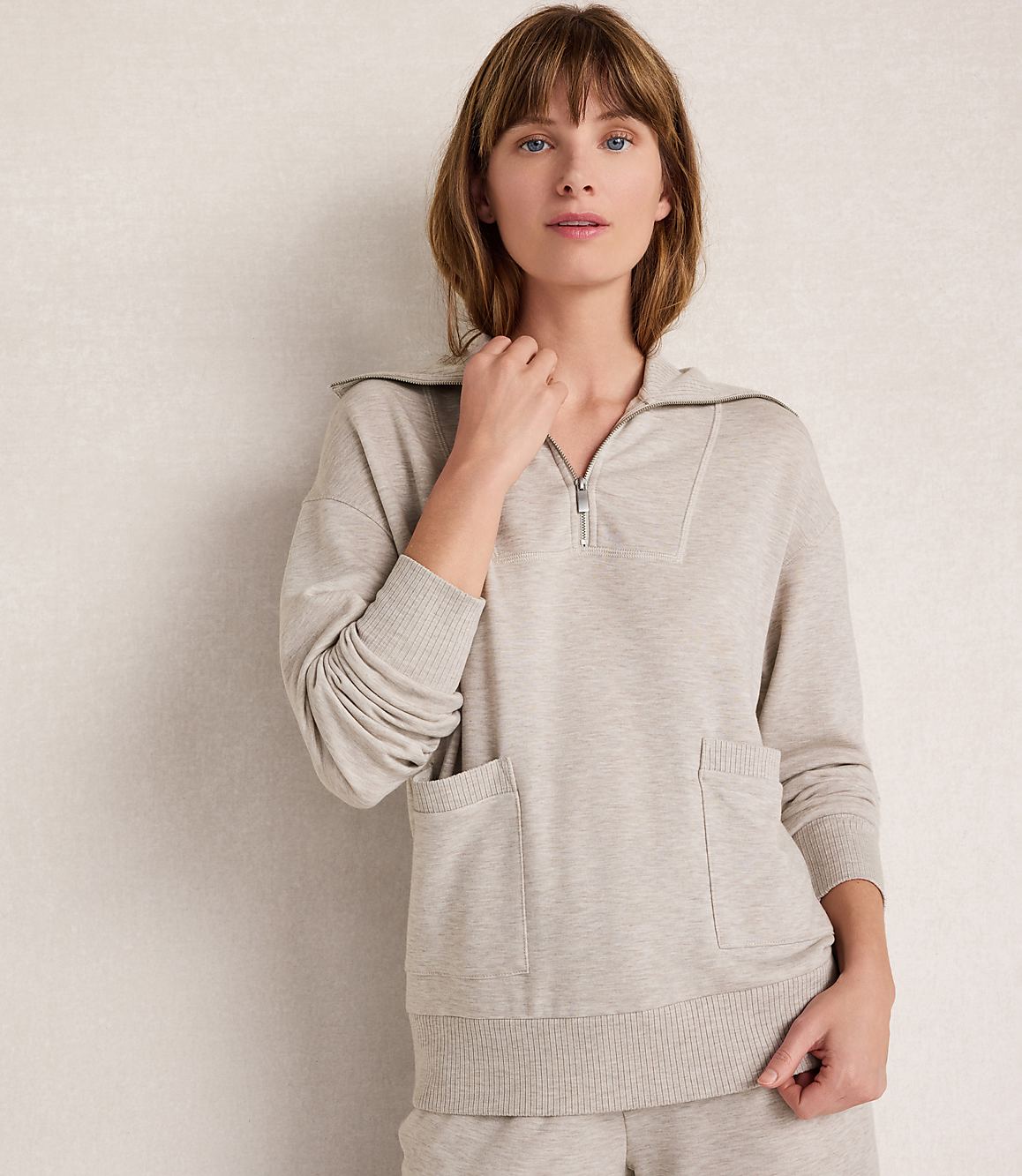 A woman in a light beige quarter-zip sweatshirt with pockets stands against a textured, light-colored wall.