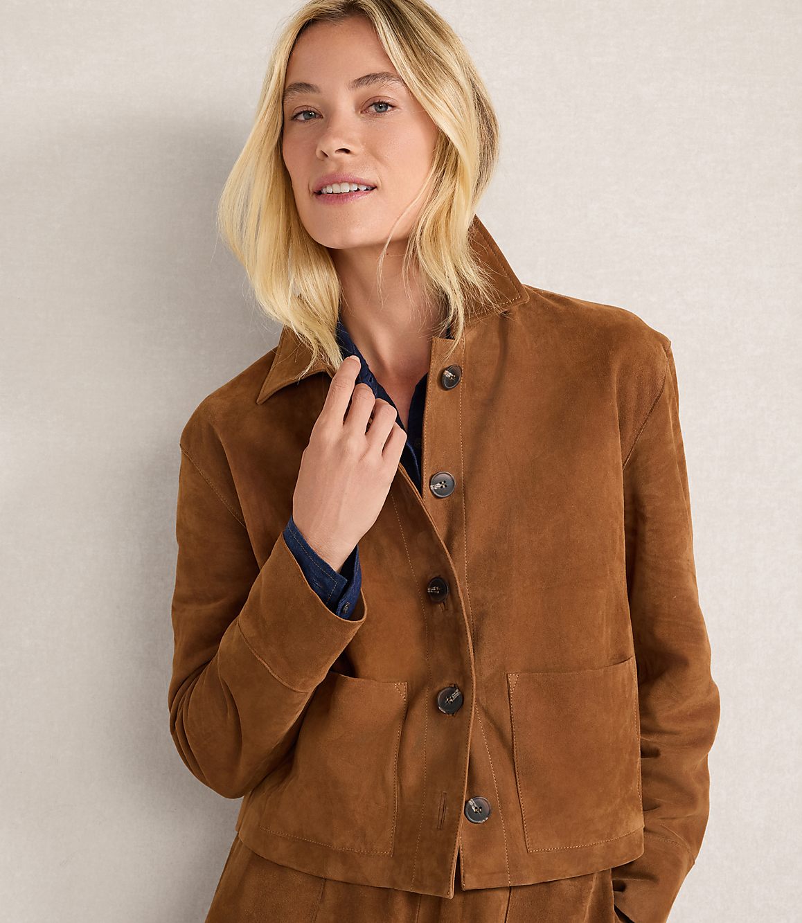 A blonde woman in a brown suede jacket stands against a neutral, textured background.