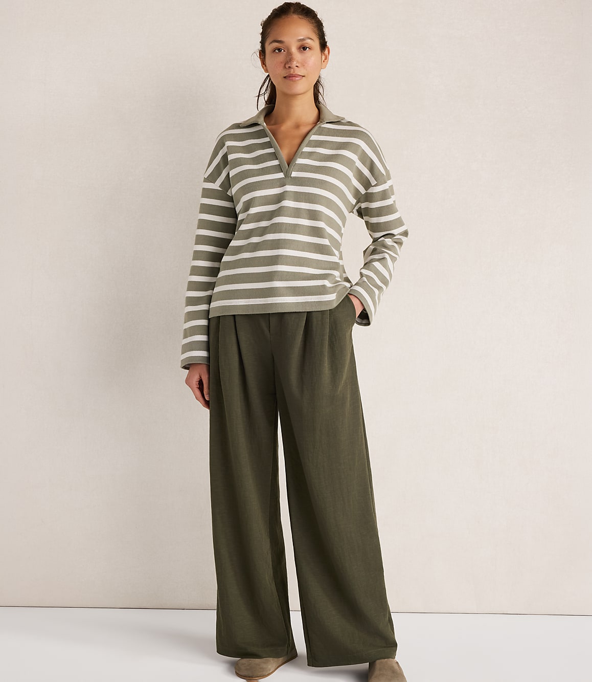 A woman in a striped collared shirt and wide-leg pants stands against a textured white background.