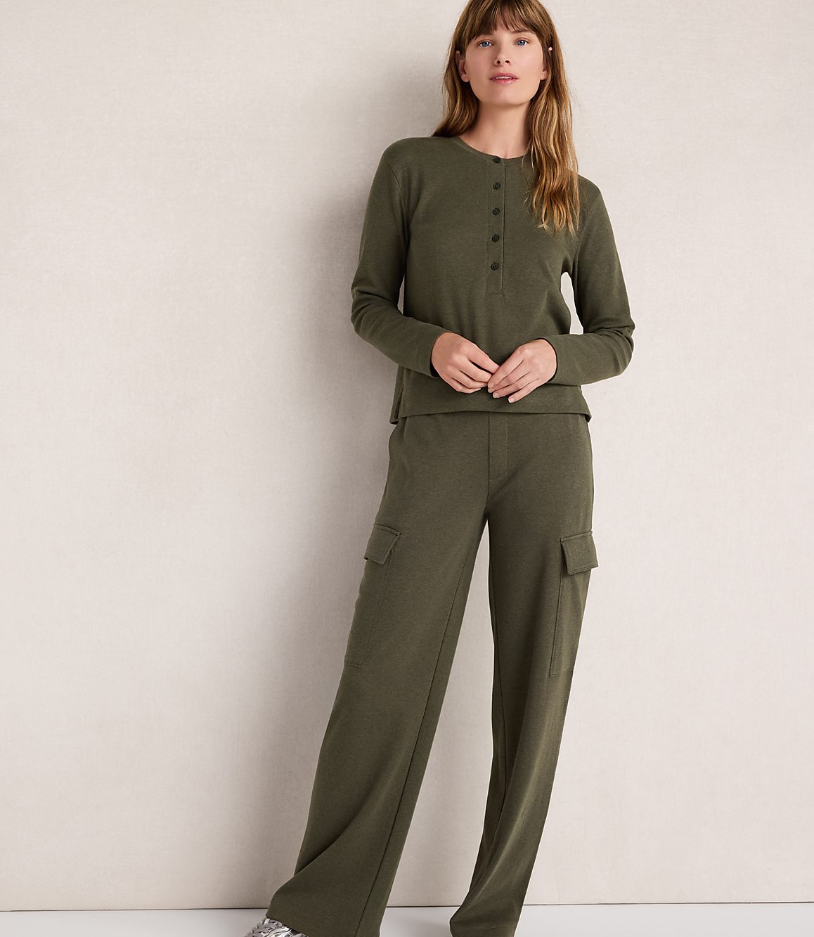 A woman in a green henley and cargo pants stands against a plain, light-colored wall.